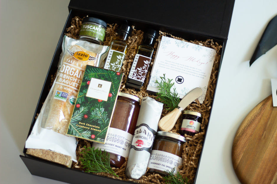  A gift basket assorted gourmet food items, including a large jar of Kansas City Canning Co. Pickled Bloody Mary Mix, a smaller jar of Rosemary Fig Spread, bottles of Olive Tree olive oil, Organic Baggett bread, Salami, Tuscany Bread Dipping Blend, and a Christopher Elbow Chocolate Bar, with a wooden cutting board and wooden spoon. 