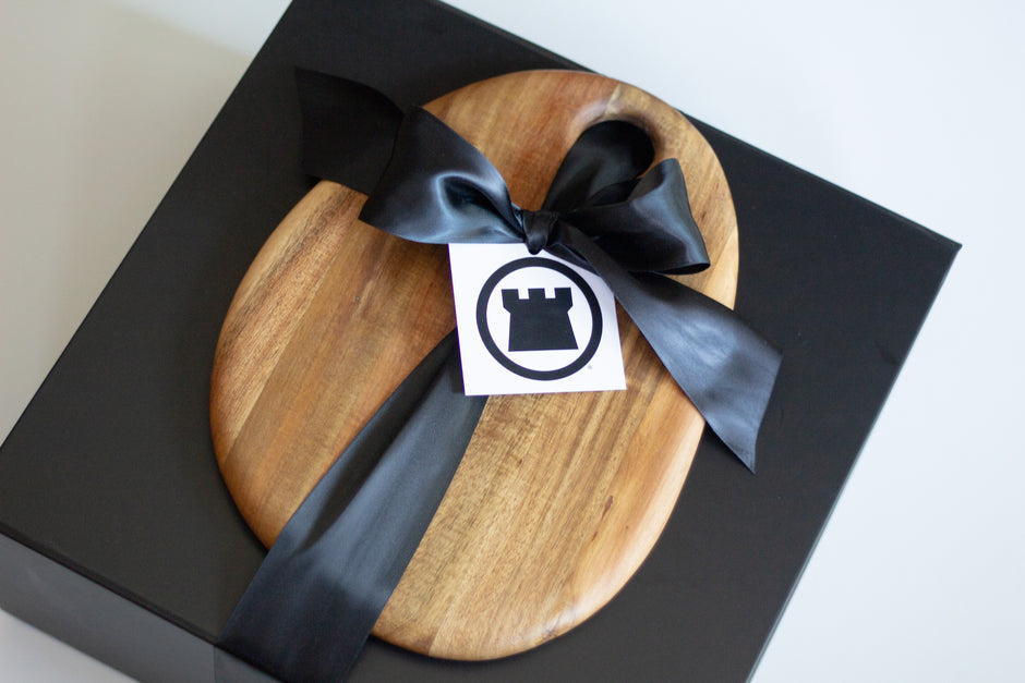 A round cutting board with a natural wood grain finish, wrapped in a black satin ribbon and tied with a black bow. A small white tag with a black castle logo is attached.