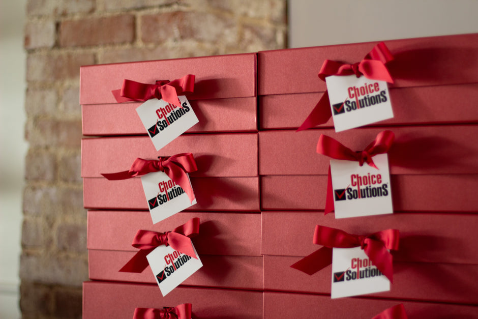 A stack of red gift boxes tied with red ribbons and custom 'Choice Solutions' hang tag, set against a rustic brick wall background.