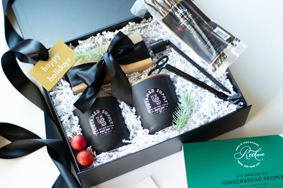 A curated gift box set featuring a black gift box filled with Untamed Candle Supply candles, a gingerbread person baking kit, and holiday decorations, alongside a green gingerbread person kit box.