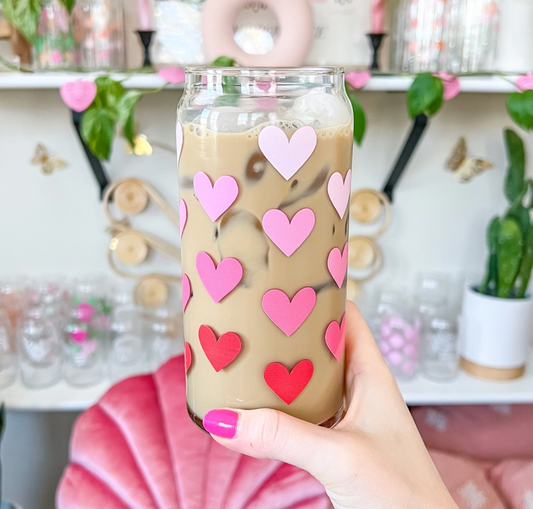 A hand with pink nail polish holds a clear glass can-shaped tumbler filled with iced coffee and decorated with pink and red heart stickers, set against a blurred background of shelves and decorative items.