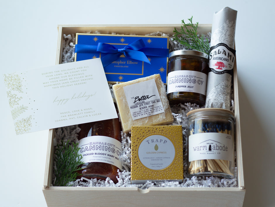A curated gift box set in a light wooden box filled with a curated gift box set from Bow On Top featuring Canning Co. Bloody Mary Mix, Pepper Jelly, Salami, Trapp Candle, Warm Abode Matches, and Christopher Elbow Chocolates. 
