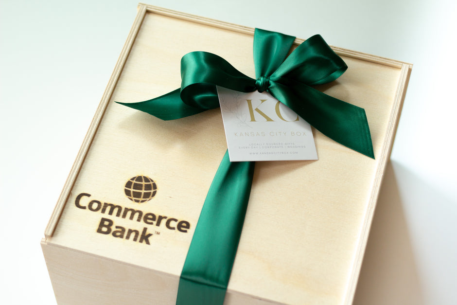 A light natural wood gift box with 'Commerce Bank' printed on the lid, tied with a dark green satin ribbon and bow, and a small white hang tag.