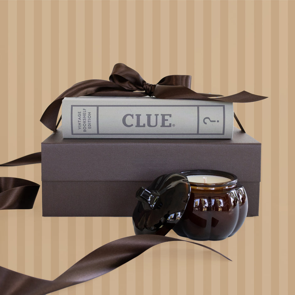 A dark brown gift box with a matching ribbon bow and a book-style board game box labeled 'CLUE.' stacked in front of a dark brown candle jar with its lid off, set against a beige striped background.