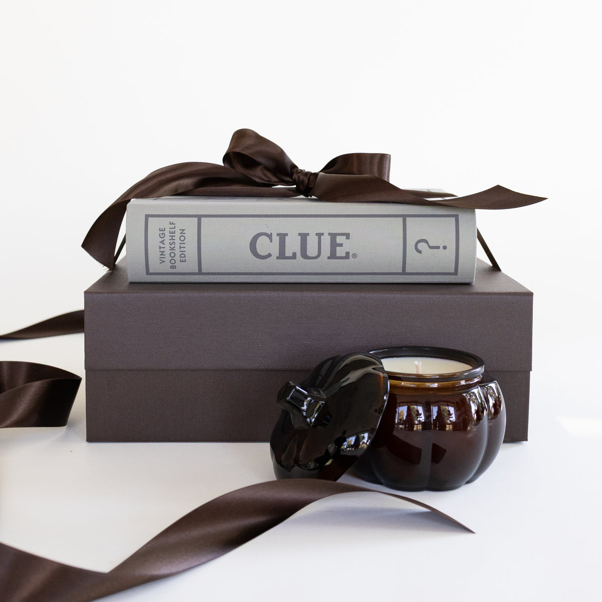 A dark brown candle in an amber glass jar with its lid off, styled on top of a dark brown gift box with a brown satin ribbon and the board game 'CLUE'.