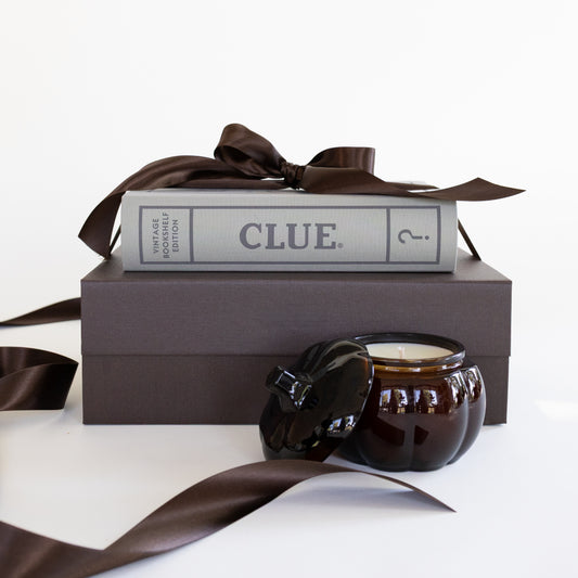 A dark brown candle in an amber glass jar with its lid off, styled on top of a dark brown gift box with a brown satin ribbon and the board game 'CLUE'.