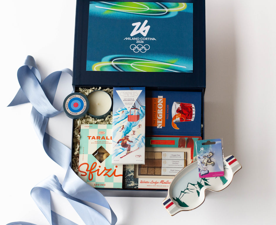 A curated flat lay of Milano Cortina 2026 Winter Olympics merchandise on a white background, complete with a light blue ribbon, a ski goggles-themed trinket dish, a blue candle in a travel tin, an Italian cocktail book, Italian crackers,  and a chocolate bar with a ski illustration on it.