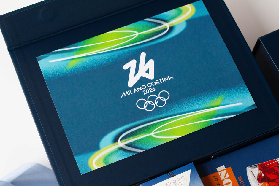 A close-up of a dark navy gift box with a printed front panel featuring the Milano Cortina 2026 Olympic logo in white over a blue abstract background with green and yellow curved accents.