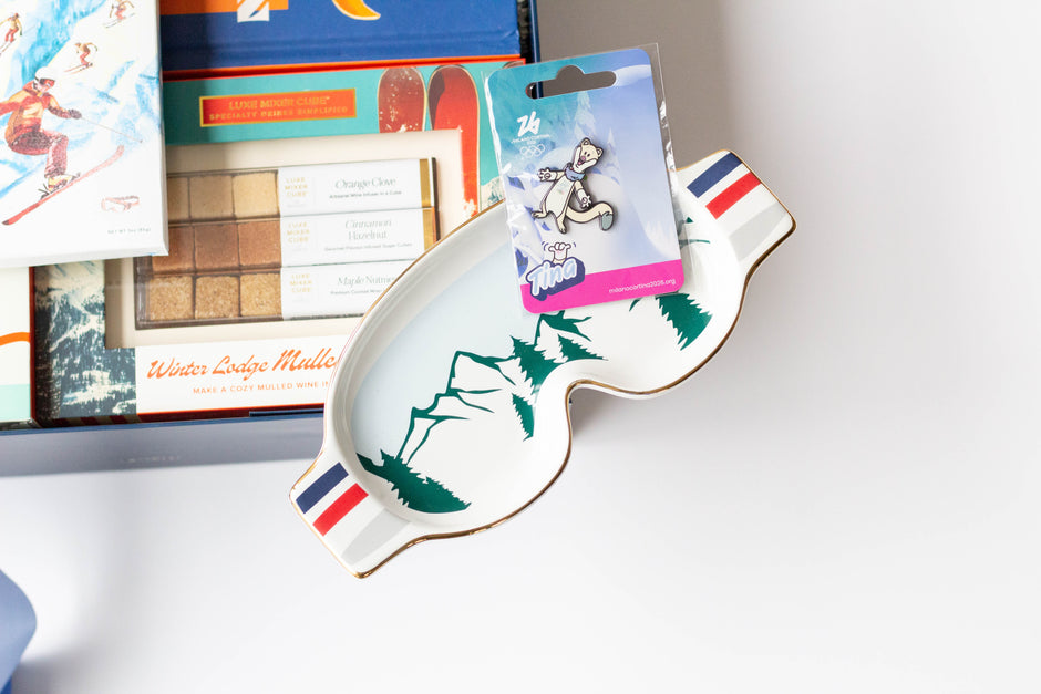 A white ceramic ski-shaped dish with green pine tree illustrations and red, white, and blue striped edges, displayed next to a gift box. A packaged metal pin featuring the Milano Cortina 2026 Olympics Mascot is resting on top of the dish.