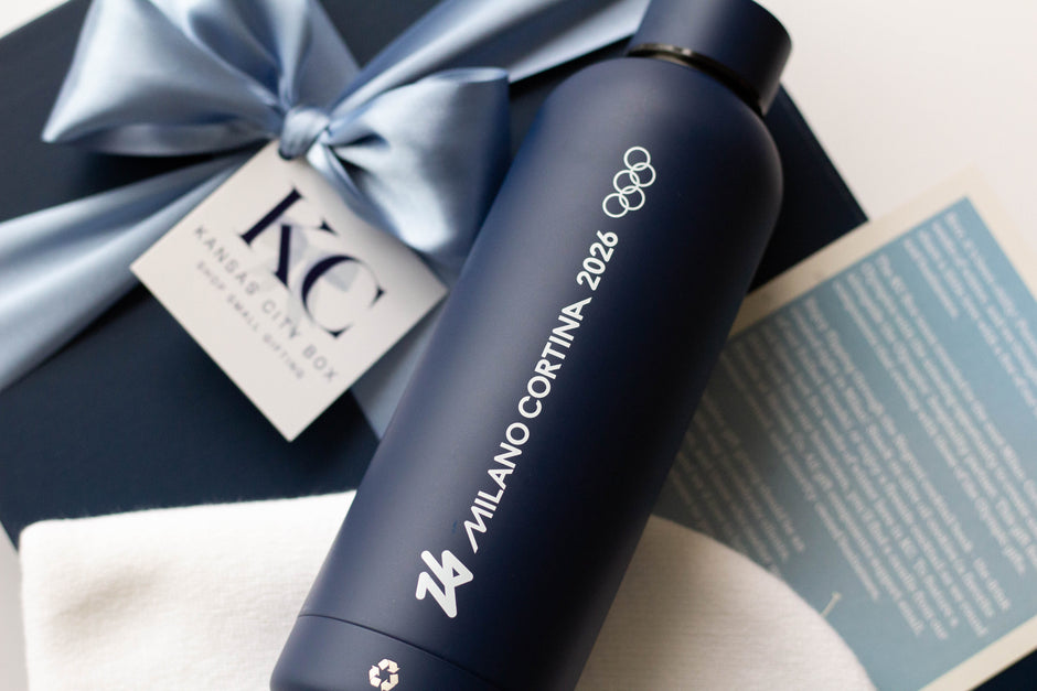 A dark navy blue cylindrical bottle with white text reading 'MILANO CORTINA 2025' and the Olympic rings logo, placed on a gift box tied with a light blue ribbon. A small card and printed insert are visible around it.