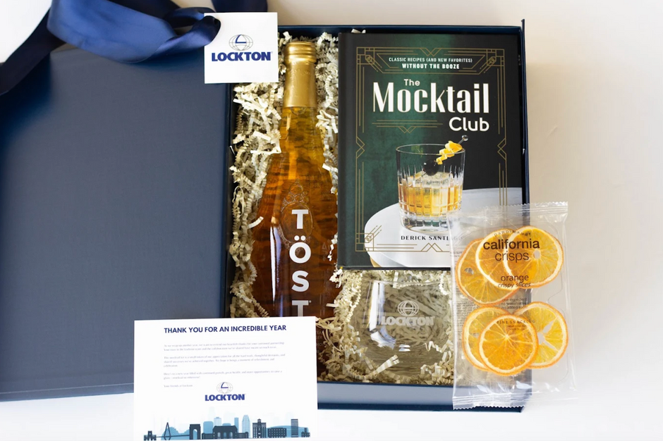 A Lockton gift box set containing a bottle of Töst non-alcoholic champagne, a book titled 'The Mocktail Club', orange slices, and a card, arranged in a black box with white shredded paper.
