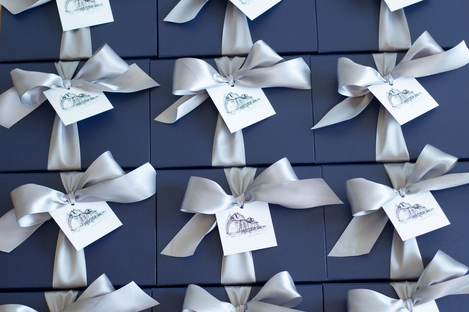 A close-up of multiple dark navy gift boxes arranged in a grid, each tied with a white satin ribbon and a small white tag printed with a delicate illustration.
