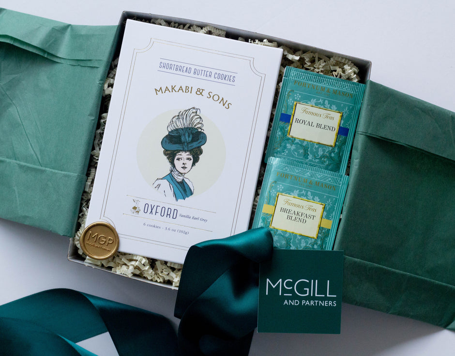 An open gift box containing a box of Makabi & Sons Oxford Butter Cookies, two packaged tea bags, a gold seal, and a dark green ribbon with a McGill and Partners tag, arranged on a light background.