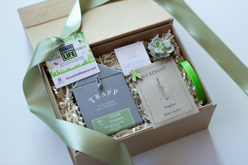 An overhead view on a plant-themed curated gift box featured a Trapp Candle, a succulent with succulent care card, Foxglove seeds from Verdant Plant Store, and a tag and bracelet from National Donate Life Month. 