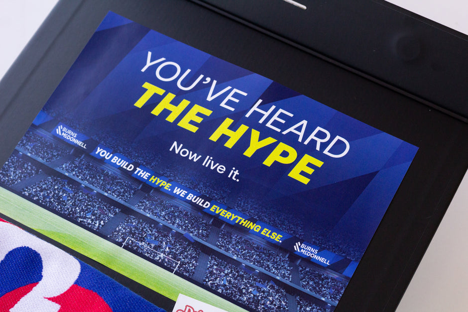 A close-up of a blue stadium graphic with the text 'YOU'VE HEARD THE HYPE' in white and yellow, plus smaller text reading 'Now live it.' and Burns & McDonnell branding.