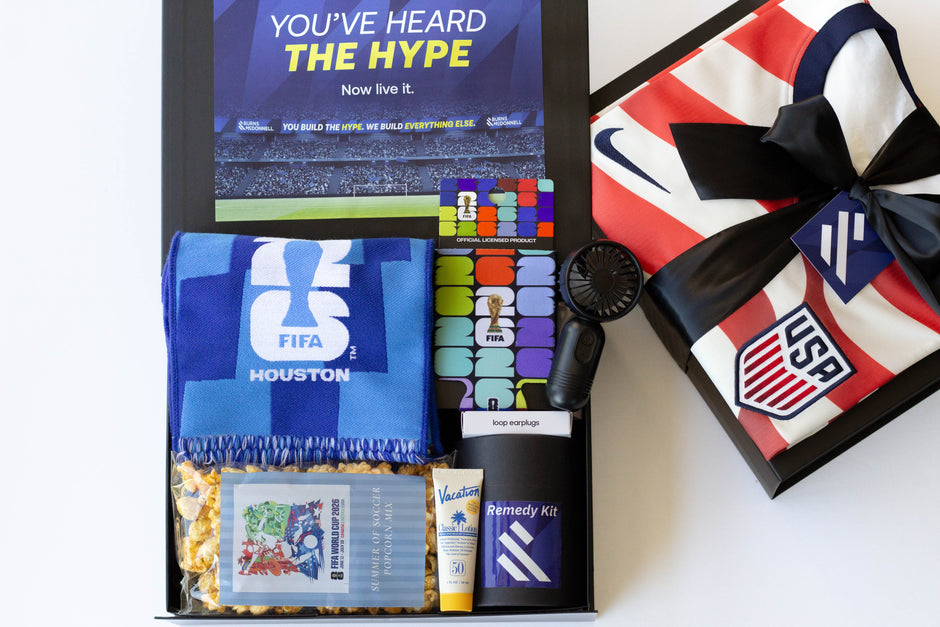 A curated gift box set featuring a blue Houston FIFA scarf, a small black portable fan, a tube of Vacation Sun Screen, a bag of popcorn, and a US Soccer Jersey on a black gift box with a black ribbon, all arranged on a white background.