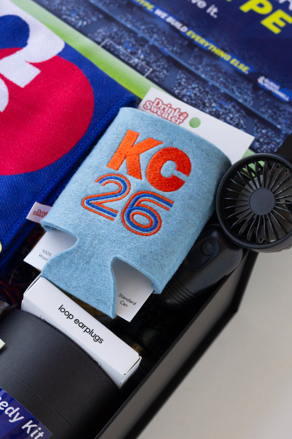 A close-up of a curated gift set containing various items, including a light blue fabric drink sweater with 'KC 26' embroidered in red and blue, a small black handheld fan, a box of loop earplugs, and other packaged goods.