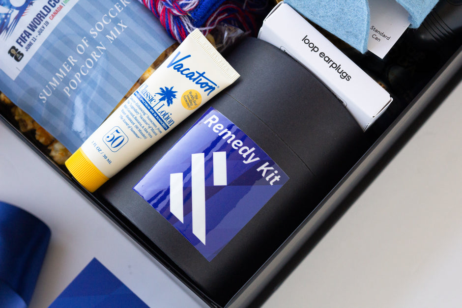 A curated wellness and travel self-care gift set arranged in a black gift box, featuring a tube of Vacation Sun Lotion, a black box labeled Remedy Kit, a box of loop earplugs, and a FIFA World Cup popcorn mix bag.