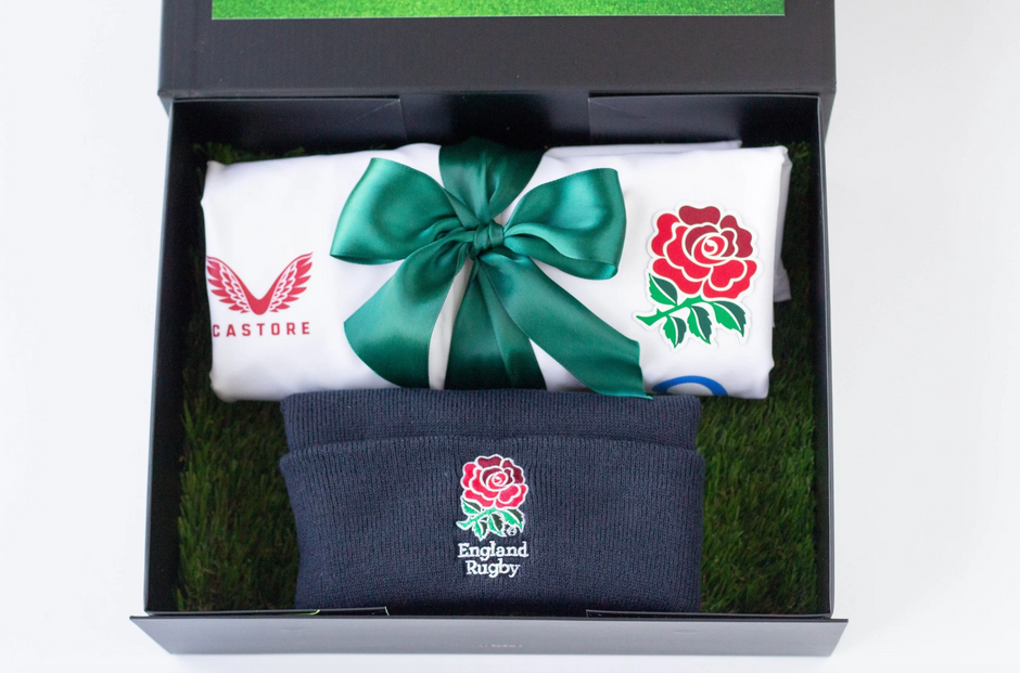 A gift set in a black box with artificial grass lining, containing a rolled white Castore rugby jersey tied with a green ribbon and a navy England Rugby knit cap with a red rose logo.