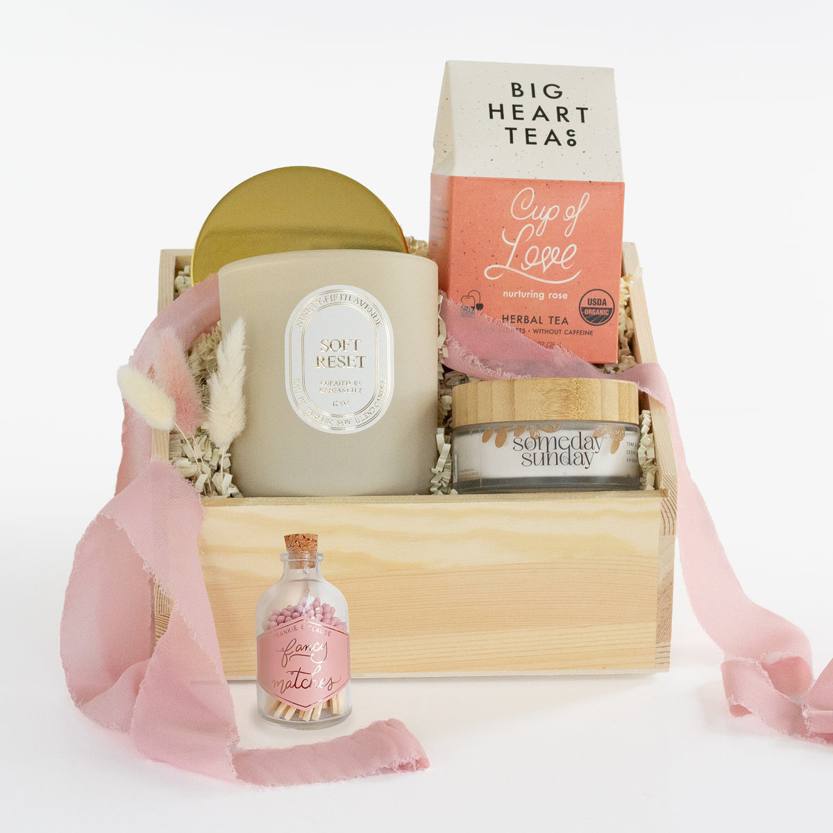 A curated gift basket set in a light wood crate with shredded paper filler, featuring a large candle, a tea box, a small jar, and a pink ribbon, photographed on a clean white background.