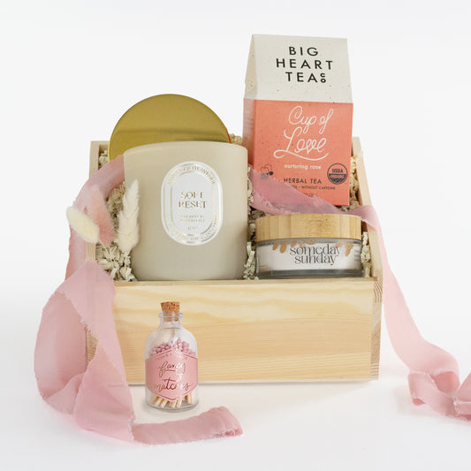 A curated gift basket set in a light wood crate with shredded paper filler, featuring a large candle, a tea box, a small jar, and a pink ribbon, photographed on a clean white background.