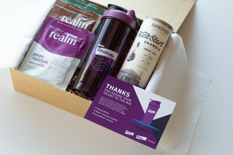 A curated wellness gift box containing a purple and white Realm smoothie mix bag, a dark purple travel tumbler with a purple lid, a white canister of Kick Start Granola, and a purple and white thank you card, all arranged inside a light wood box.