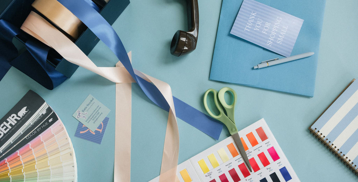 A styled flat-lay of craft and design supplies on a light blue surface, including a spool of blue ribbon, scissors, a color swatch fan, a notebook, and other stationery items.