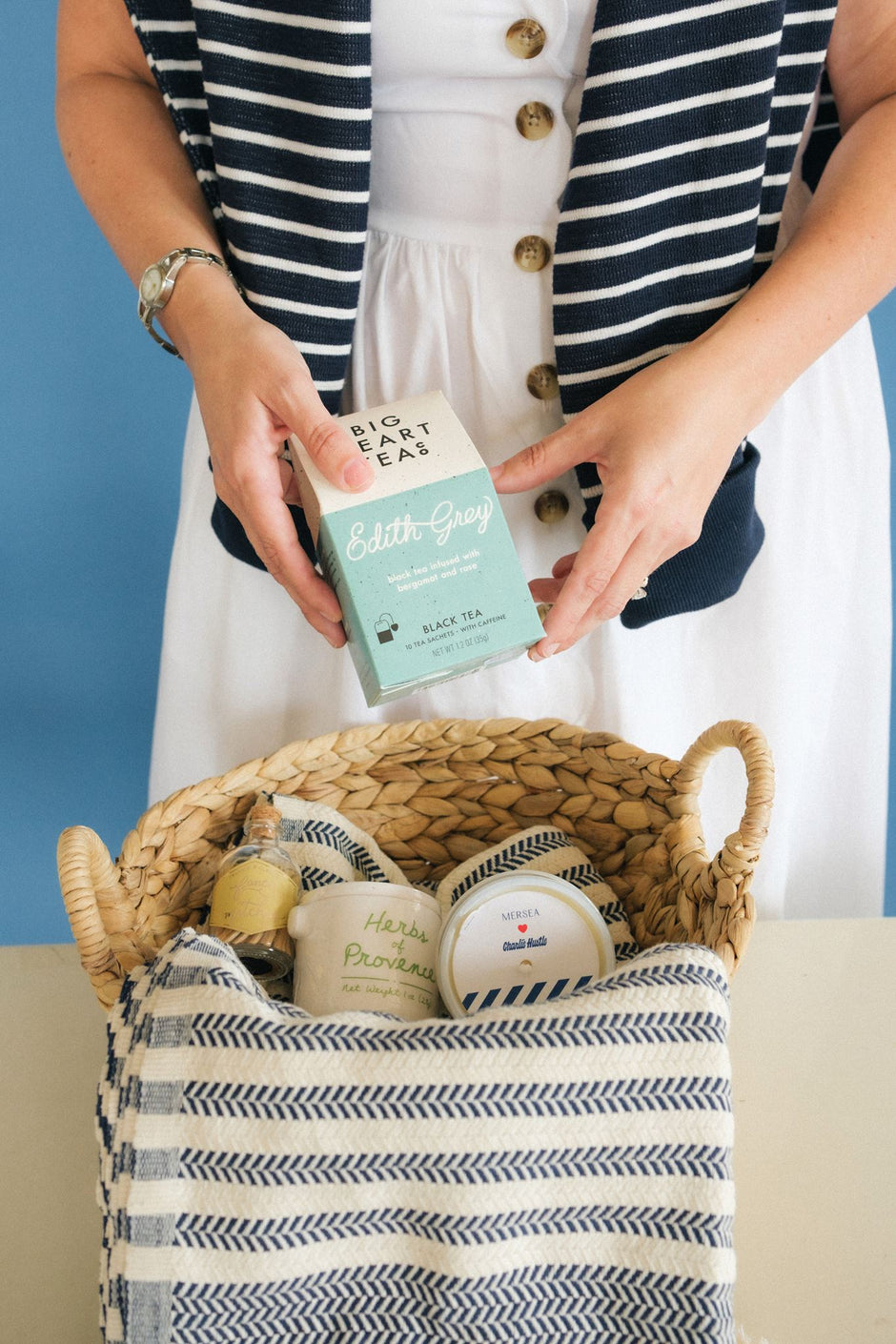 A person in a navy and white striped cardigan over a white dress holds a tea box above a woven basket containing home decor gifts like matches, herbs, and a candle, against a blue background