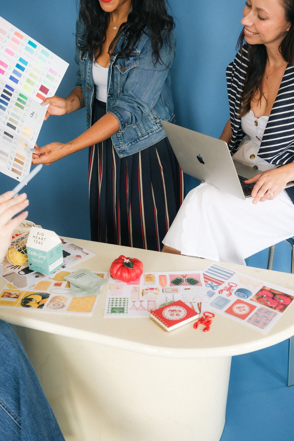 Three women collaborating around a white table filled with colorful printed cards, swatches, and small stationery items against a blue wall. One woman holds a large color chart, another uses a laptop, and the table displays a variety of printed designs.