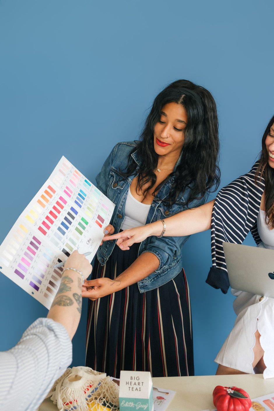 Three women gathered around a color swatch chart in a bright room with a blue wall. One woman holds the chart while another points at it, and a laptop is visible on the table.