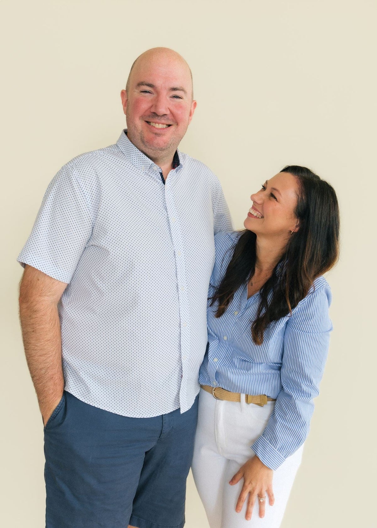 A smiling couple posing together against a solid light beige background. The man on the left is bald and wearing a light blue and white micro-checkered short-sleeve button-down shirt with navy blue shorts.