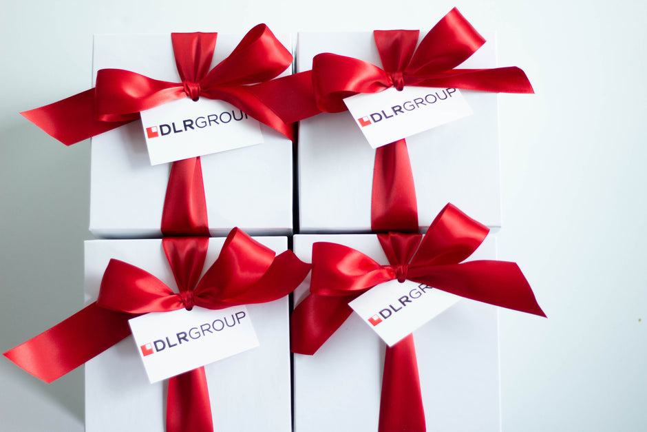 Four white gift boxes arranged in a 2x2 grid, each tied with a large red satin ribbon and a small white tag printed with the brand name DLRGROUP.