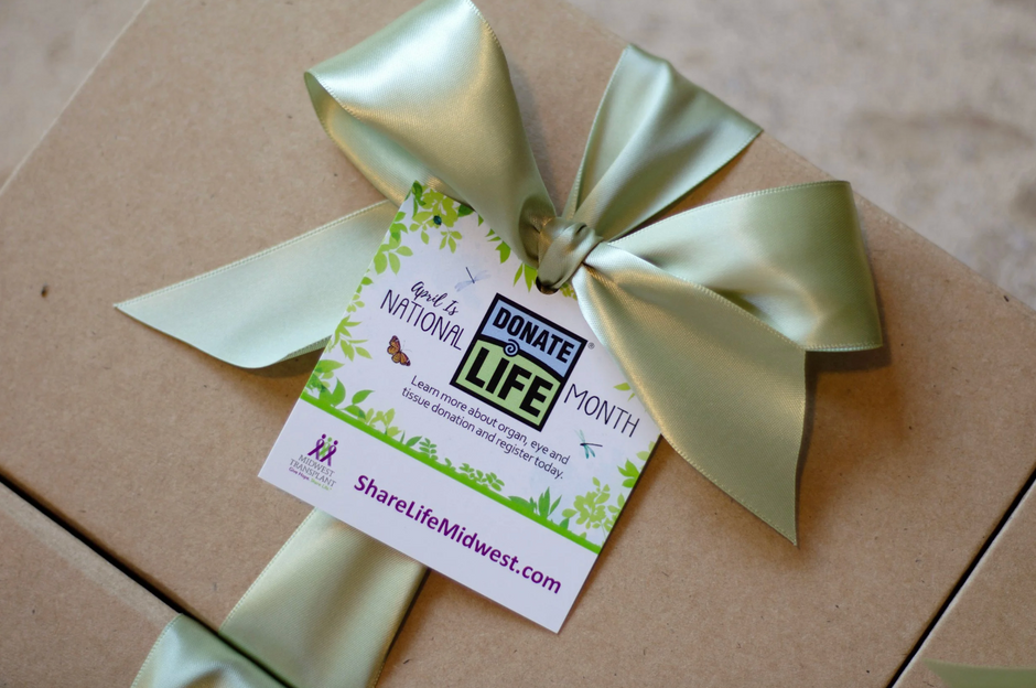 A close-up of a kraft paper gift box tied with a large pale green satin ribbon and a hanging tag that reads 'Donate Life Month' and 'ShareLifeMidwest.com'.