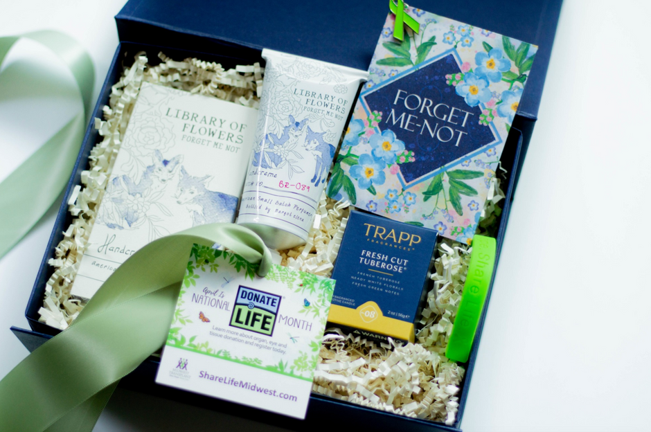 A curated gift box set containing a 'Forget Me Not' floral box, a 'Library of Flowers' book, a tube of hand cream, a Trapp Fresh Tuberose Candle, and a Donate Life Month card, arranged in white crinkle paper inside a navy blue box with a green ribbon.