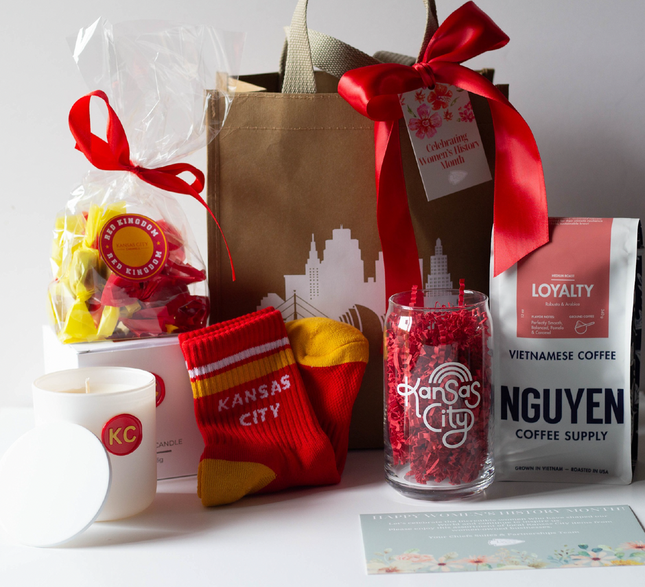 A curated gift set featuring a brown paper bag with a red bow, a bag of yellow and red wrapped caramels, a white candle with a red KC logo, a pair of red and yellow Kansas City socks, a beer glass tumbler that reads "Kansas City", and a bag of Nguyen Vietnamese Coffee Supply Loyalty coffee.