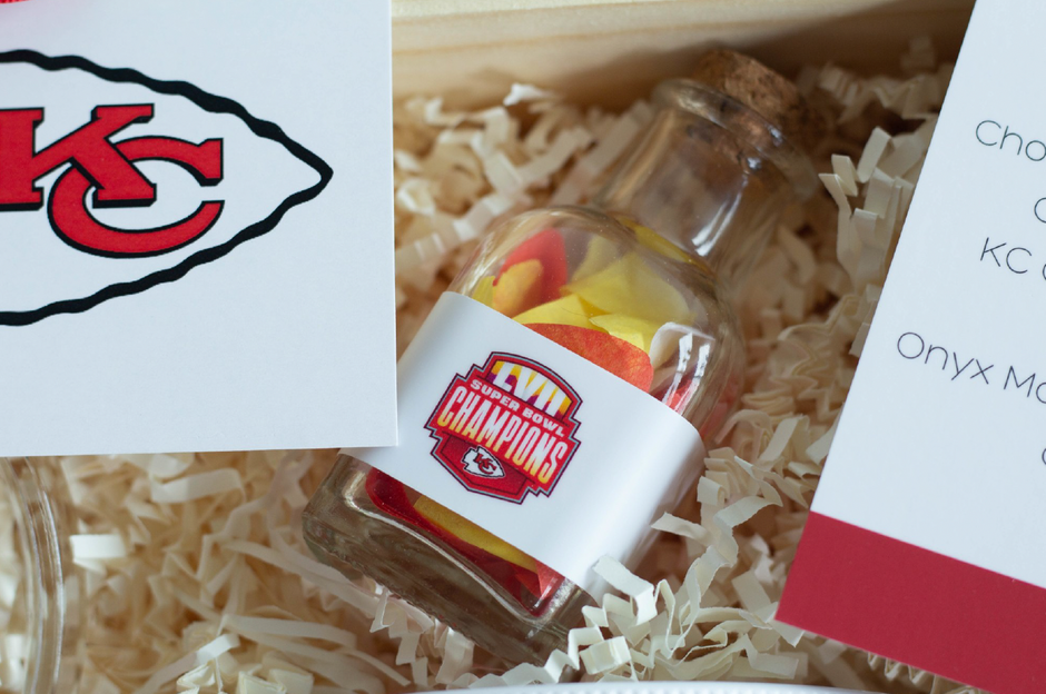 A small clear glass bottle with a cork stopper sits in shredded paper packaging. The bottle has a white label featuring the Kansas City Chiefs logo and the word 'CHAMPIONS.