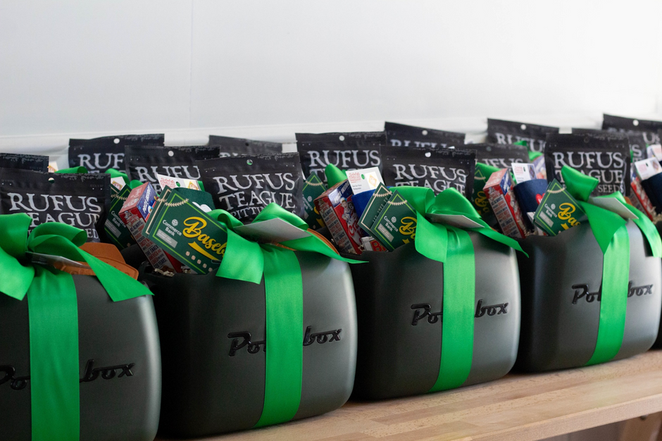 A row of black Polarbox Coolers as gift baskets tied with bright green ribbons, filled with packaged snacks and drinks, including Rufus Teague sunflower seeds, arranged on a wooden shelf against a plain white wall.