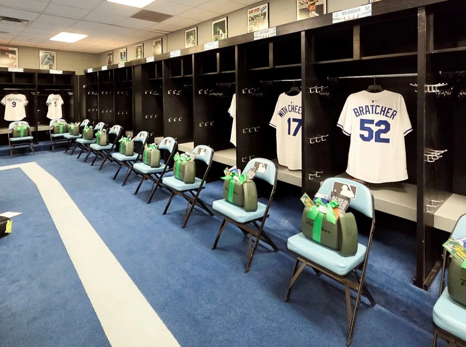 A baseball team locker room with dark wood lockers, white jerseys hanging in the cubicles, folding chairs with black and green gift boxes placed on them, and blue carpeting with a white foul-line style stripe.