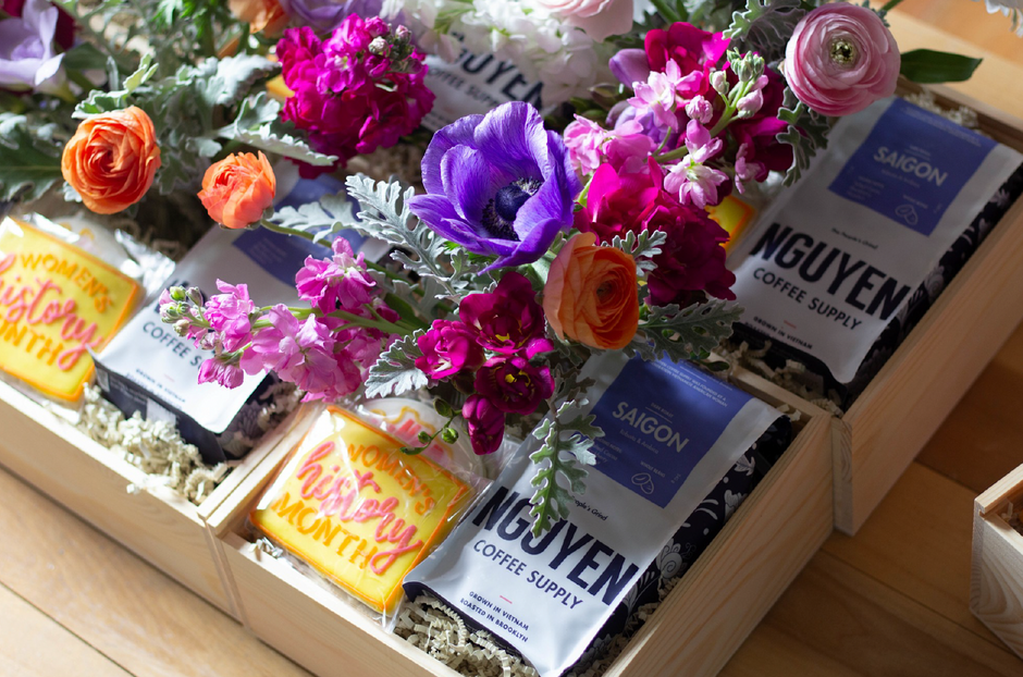 A gift box arrangement featuring bags of Saigon Nguyen Coffee Supply coffee nestled in decorative flowers and greenery inside a wooden crate.
