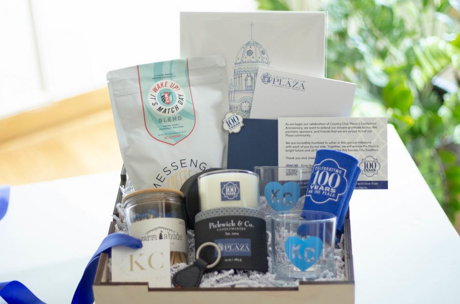 A curated gift box containing a bag of Messenger coffee, a candle, two small clear tumblers with blue heart designs, a keychain, and printed stationary, arranged in white paper shred inside a light box with a blue ribbon.