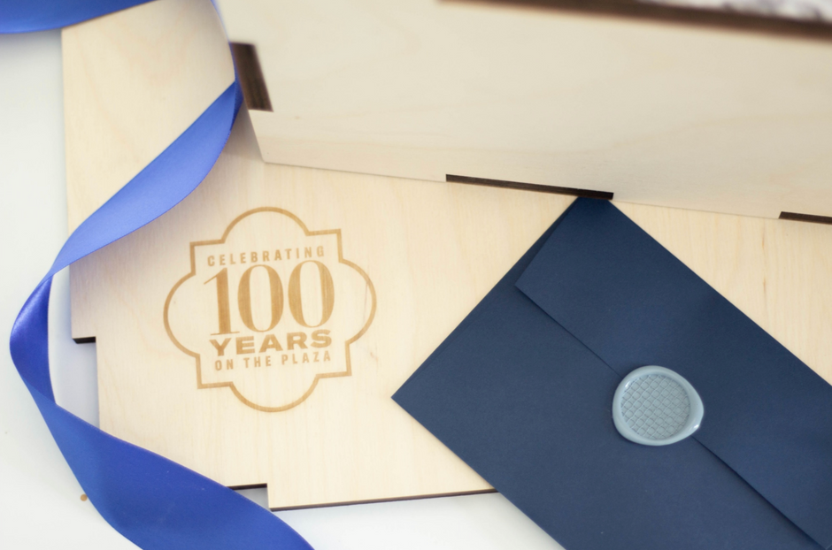 A close-up of a wooden gift box lid engraved with 'Celebrating 100 Years On The Plaza,' alongside a navy blue envelope with a silver wax seal and a blue ribbon.