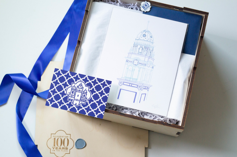 An open gift box containing a white art print with a blue architectural line drawing, a blue ribbon, a blue patterned tag, and a wooden box top with a '100 Years' emblem, photographed on a light neutral background.