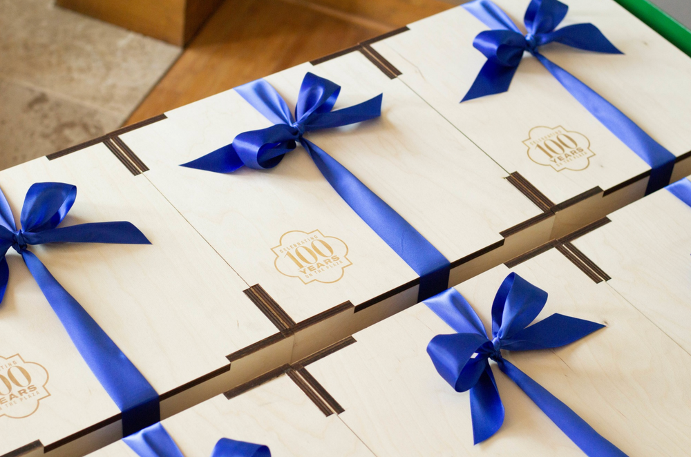 A close-up of several wooden gift boxes arranged diagonally, each tied with a bright blue satin ribbon and bow. A small gold crest logo is printed on the top of the boxes.