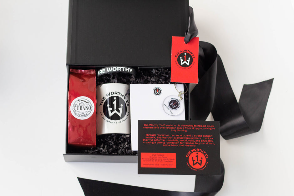 An open black gift box on a white background containing a red bag of coffee from Cafe cubano, a white mug with a black logo, a white notepad, and a black gift insert with red text. A black ribbon and red hang tag with a crown logo are also visible.
