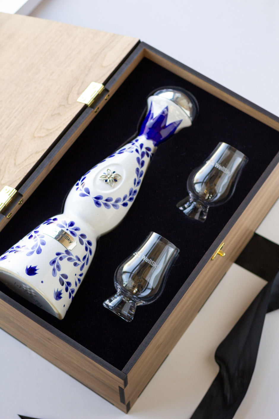 A white ceramic tequila bottle with blue floral patterns, presented in a wooden gift box with two clear glass tumblers on a black felt lining.