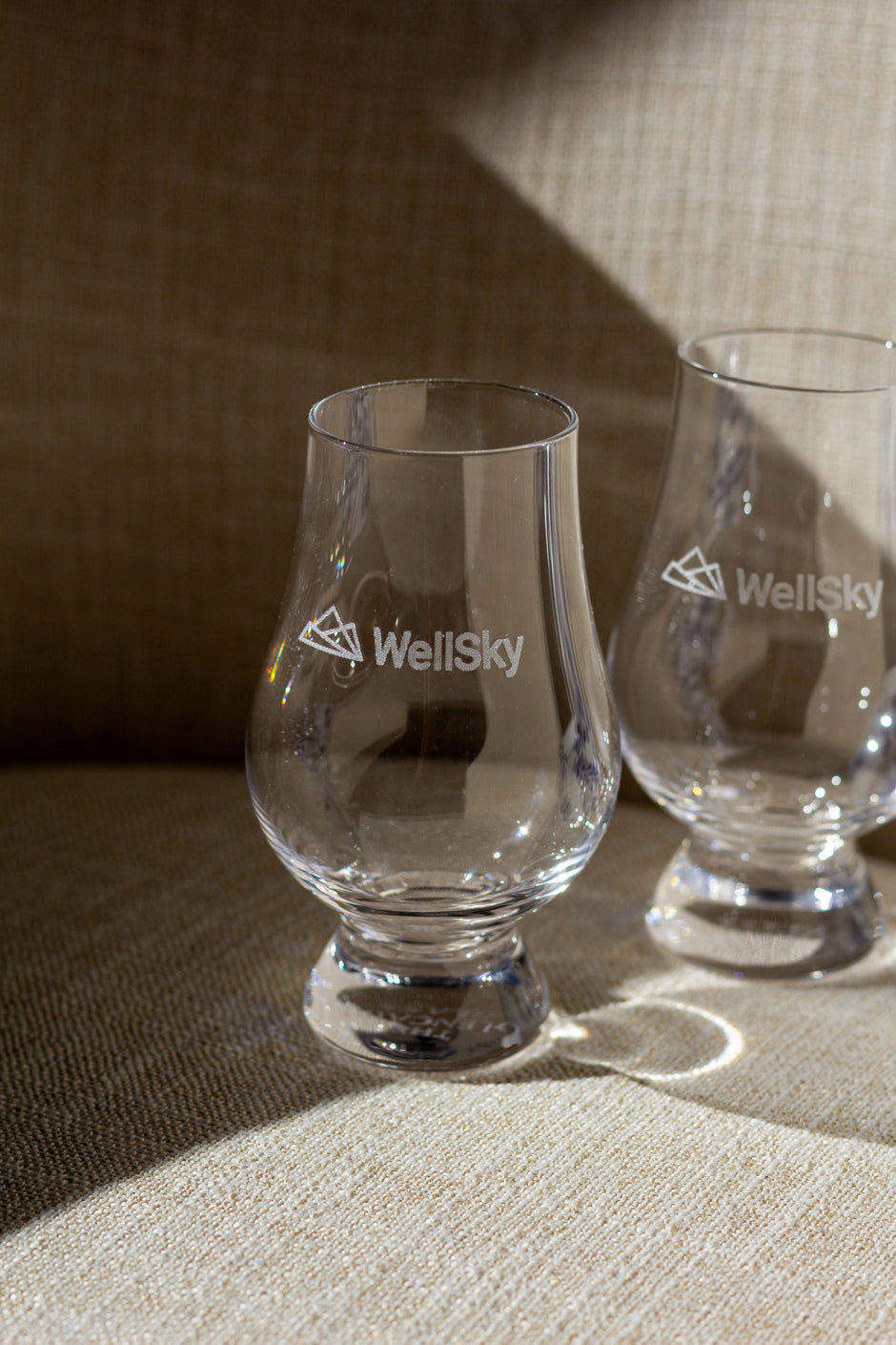 Two whiskey snifter glasses branded with WellSky logos sitting in harsh sunlight in a beige textured armchair. 