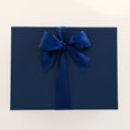 Load image into Gallery viewer, A dark navy blue rectangular gift box with a large matching satin bow on top, photographed against a plain off-white background.
