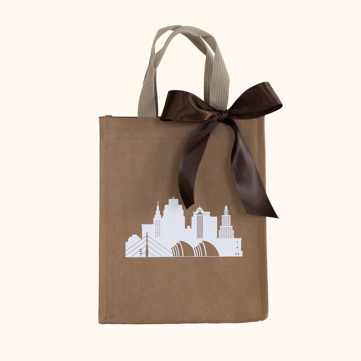 A reusable brown gift tote with beige handles, decorated with a white Kansas City skyline graphic and a large dark brown satin bow, set against a solid cream background.
