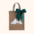 Load image into Gallery viewer, A reusable brown gift tote with beige handles, decorated with a white Kansas City skyline graphic and a large emerald green satin bow, set against a solid cream background.
