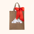 Load image into Gallery viewer, A reusable brown gift tote with beige handles, decorated with a white Kansas City skyline graphic and a large red satin bow tied around the top, shown on a plain light beige background.
